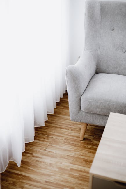 A white, upholstered sofa with tufted back and curved armrests is positioned against a light grey wall decorated with elegant, molded paneling. The sofa is placed on a wooden floor with a natural fini